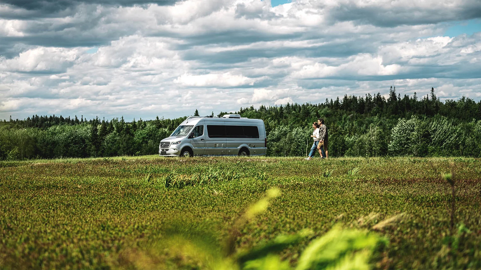 A couple walks beside a parked camper van in a vast grassy field, with a forest and cloudy sky in the background.