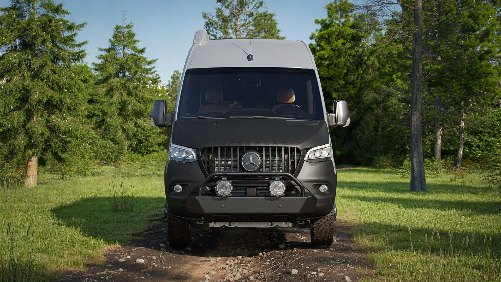 A grey off-road van parked on a dirt path in a forested area, surrounded by green trees and grass under a clear sky.