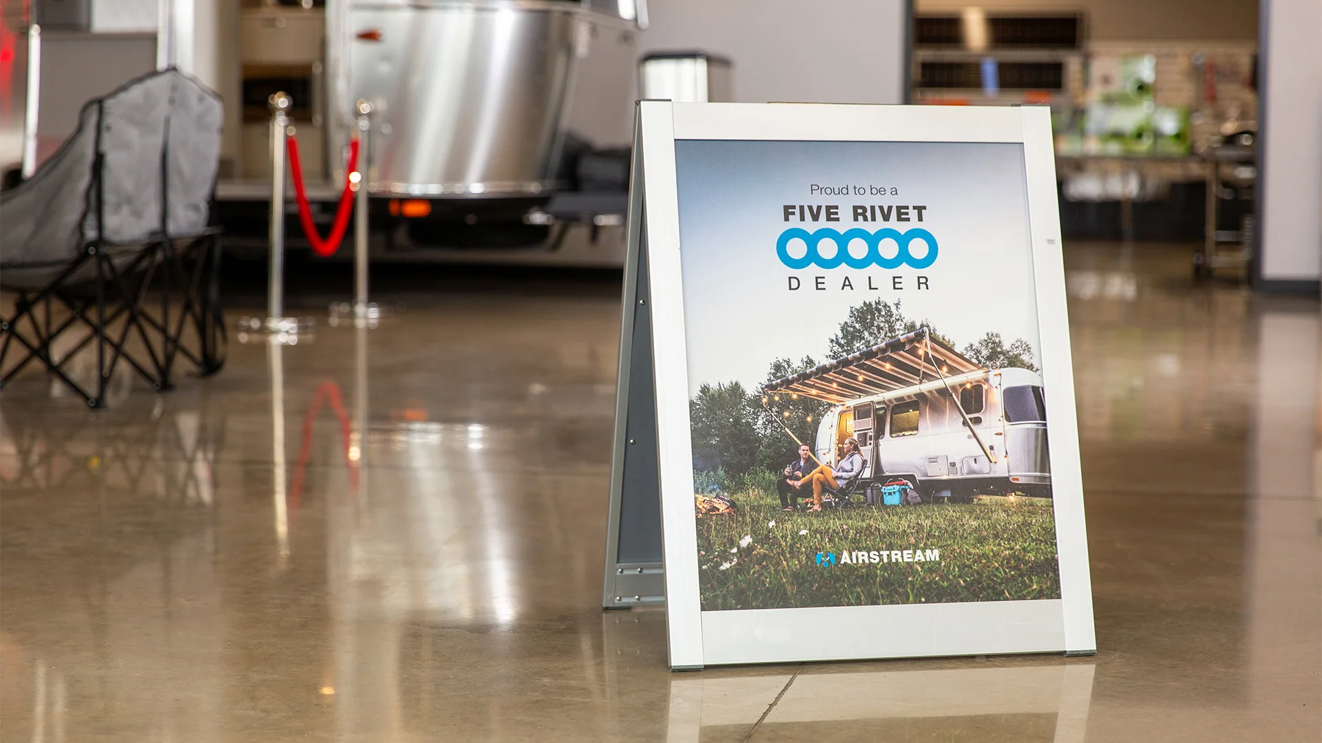 A sign on a showroom floor reads "Proud to be a Five Rivet Dealer" with an image of a camper and people outdoors, promoting Airstream.