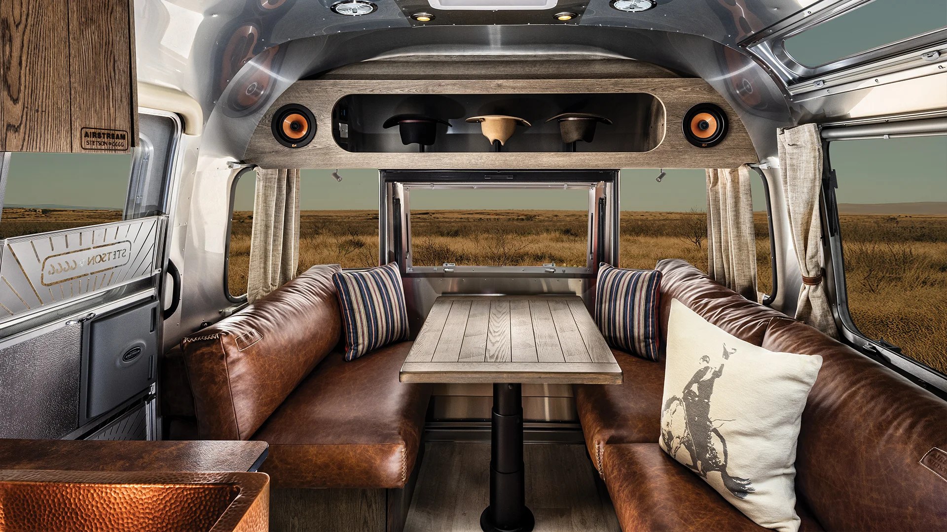 Interior of a modern campervan with brown leather seating, a wooden table, striped pillow, and a window view of a grassy landscape.