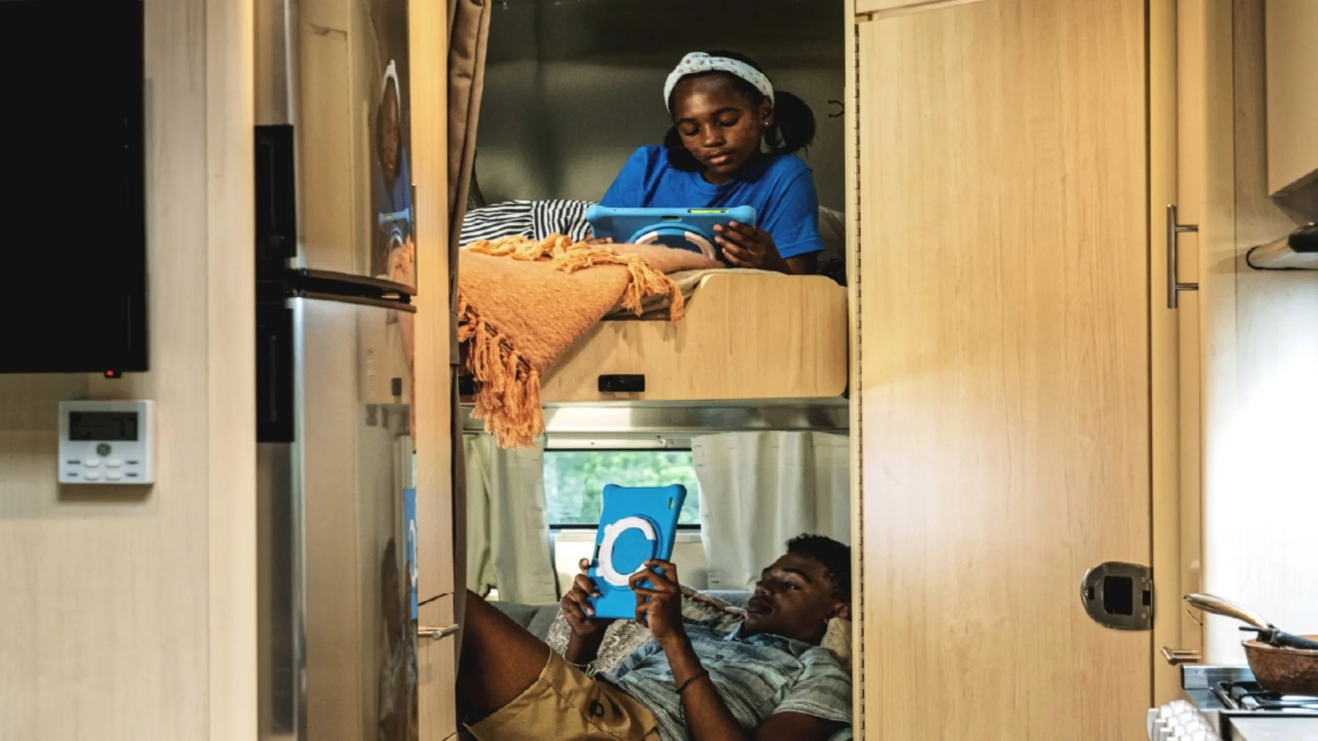 Two people in a cozy camper van relax on bunk beds, each absorbed in using a tablet, with a soft light filtering through curtains.