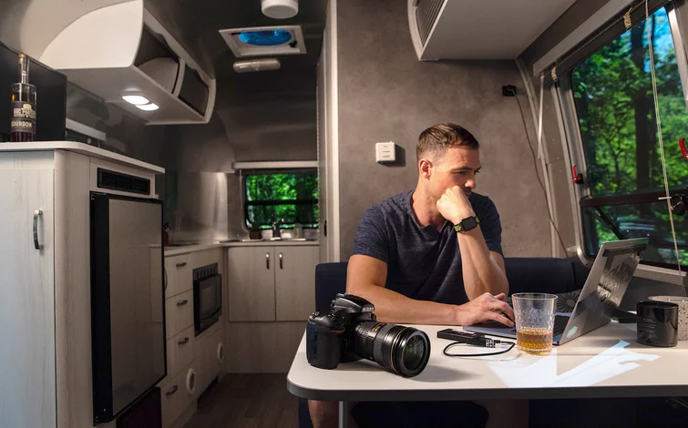 Man working on a laptop inside a camper van, with a camera and a drink on the table, surrounded by a modern kitchenette and large windows.