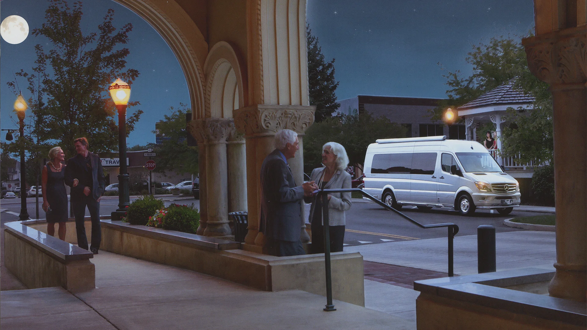 People chatting outside a building under an arched entrance at night; a white van is parked nearby under a starry sky.