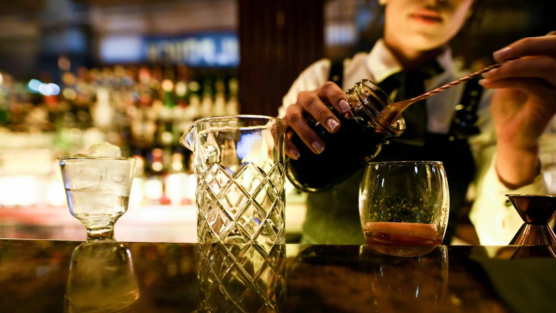 Bartender preparing a cocktail with ice-filled glasses on the bar, using a mixing spoon and pouring from a shaker in a dimly lit setting.