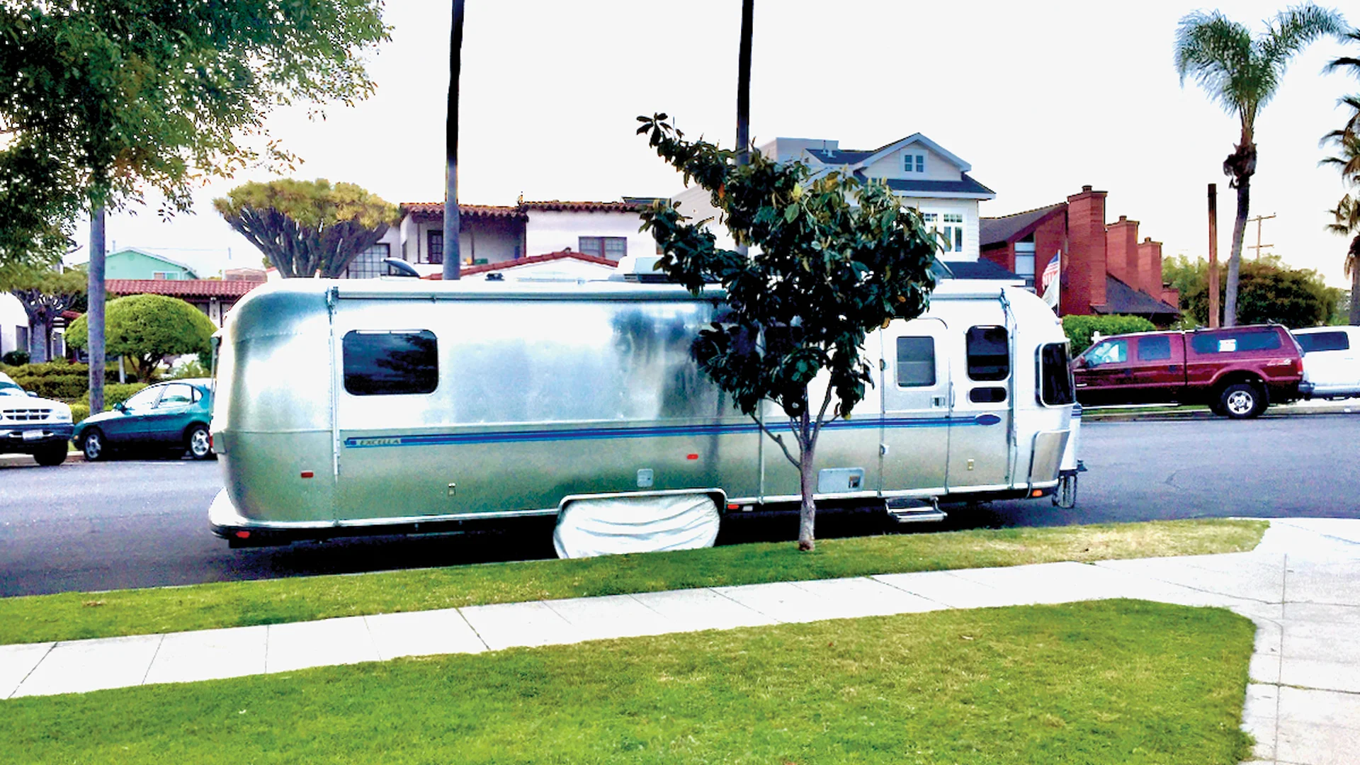 A silver Airstream trailer is parked on a residential street lined with houses, trees, and parked cars. A small tree partially blocks the view of the trailer. Grass and sidewalk are in the foreground.