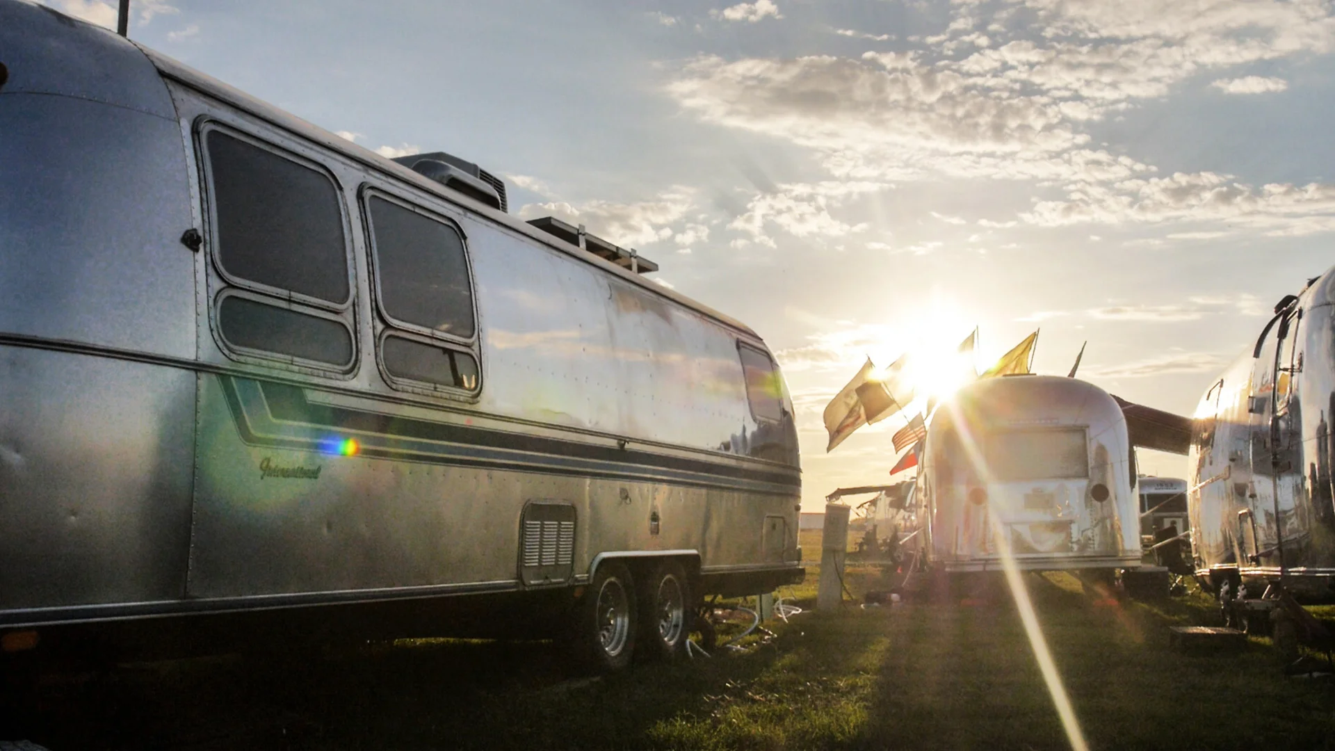 Shiny Airstream trailers parked outdoors with sunlight streaming through, flags waving, and a blue sky with scattered clouds above.