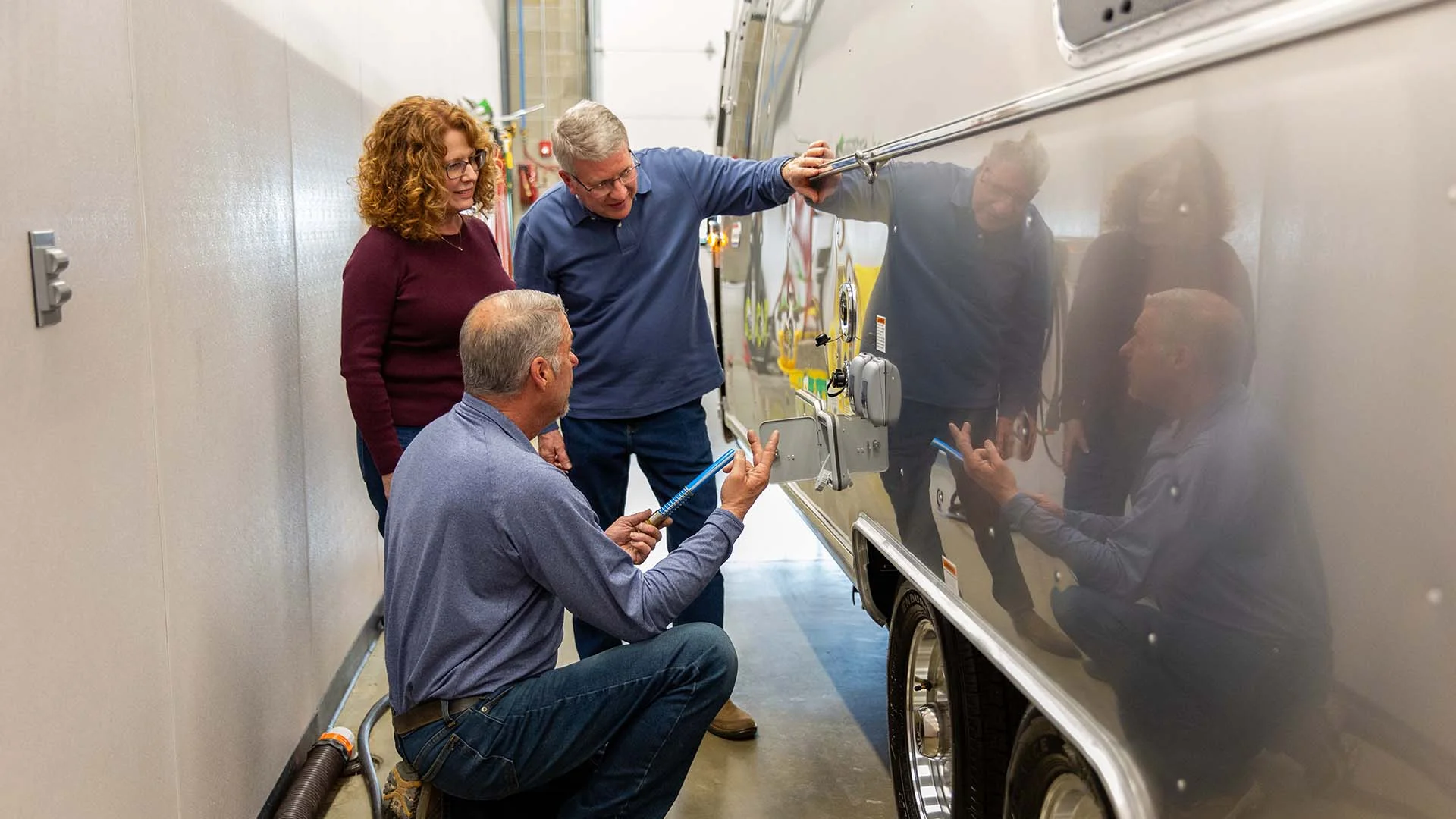 Three people inspect a shiny metallic trailer in a garage, with one person kneeling and pointing at a section of the trailer's exterior.