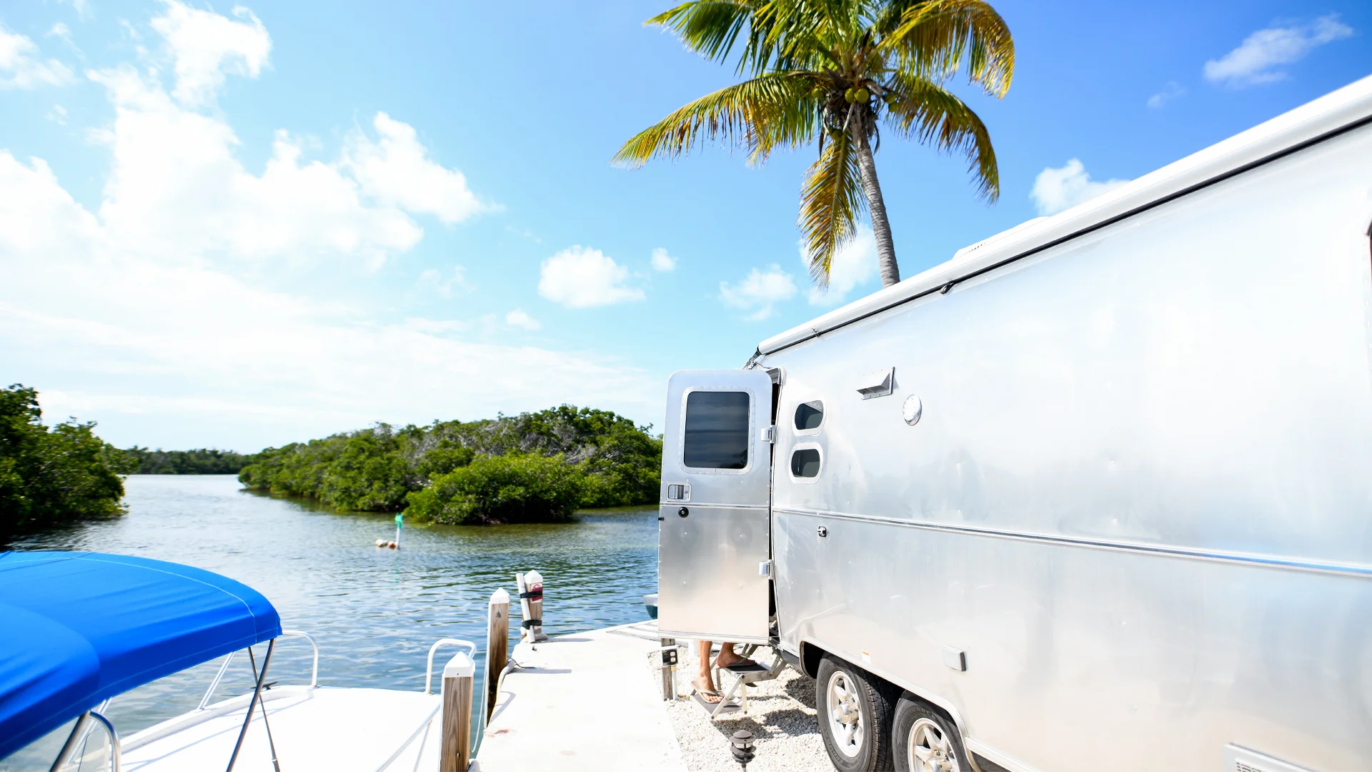 Silver camper trailer parked by a peaceful waterfront, with a palm tree overhead and blue sky filled with scattered clouds.