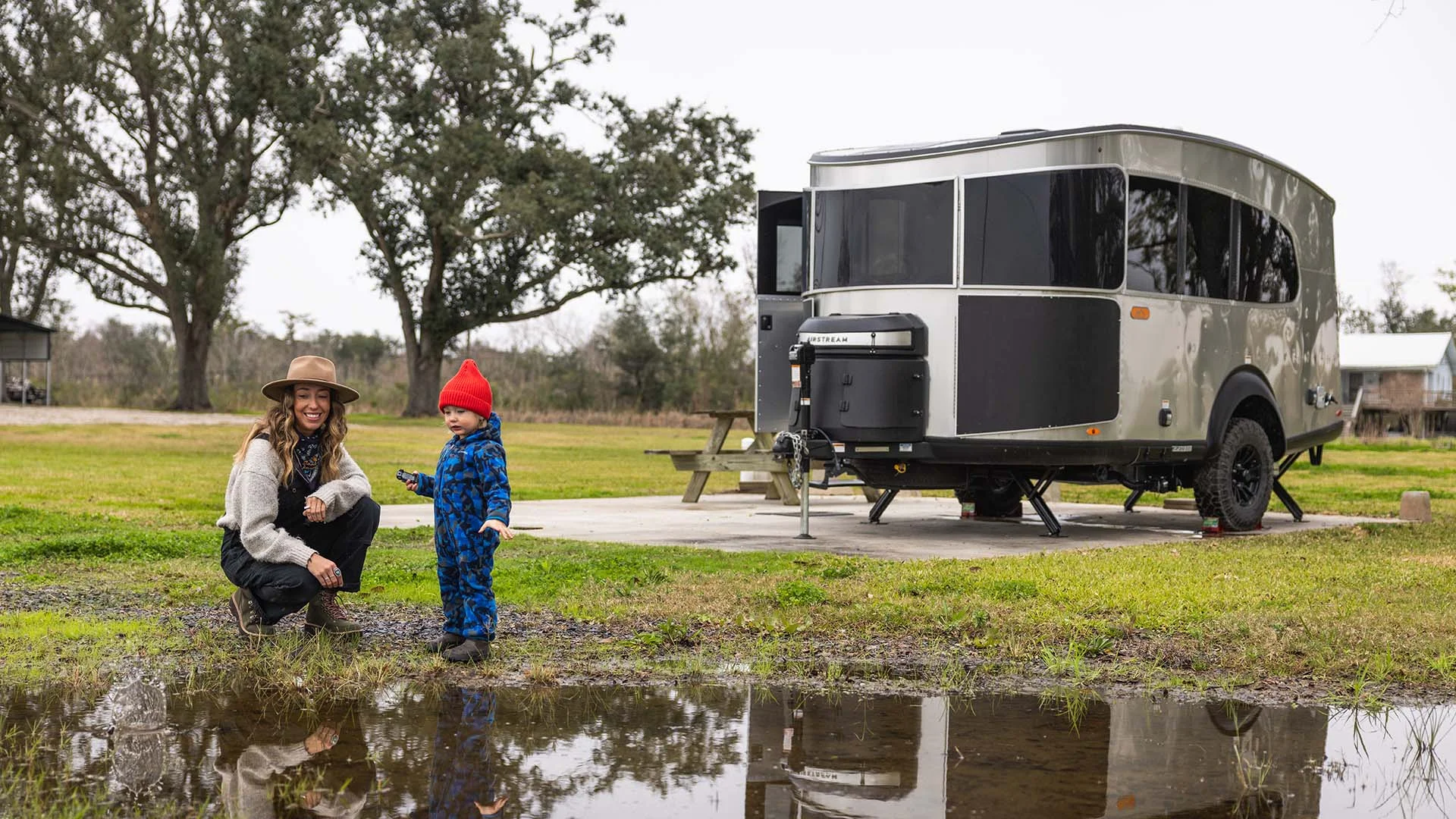 A woman and a child in a red hat stand near a puddle, with a modern silver trailer and trees in the background.