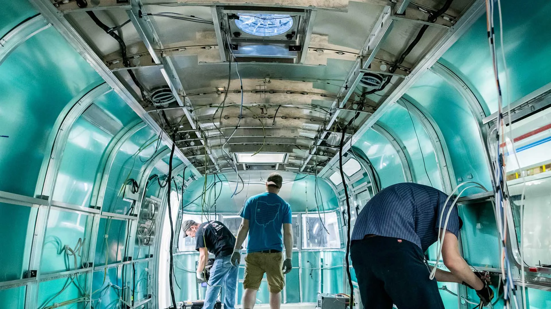 Workers installing wiring inside an unfinished, metallic trailer interior with exposed walls and ceiling panels.