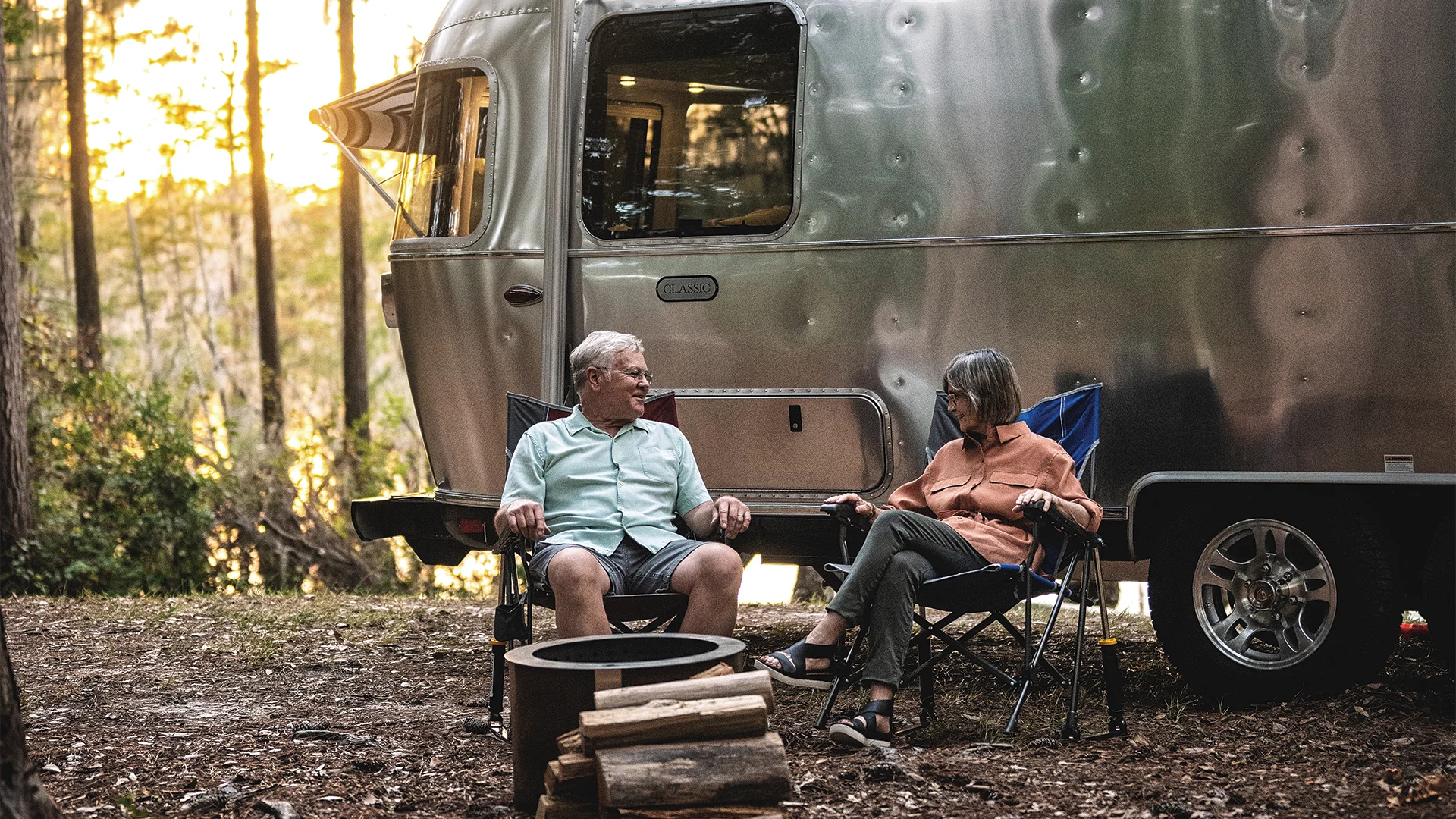 An older couple sits in camping chairs beside a silver trailer in a wooded area, with a small campfire pit nearby.