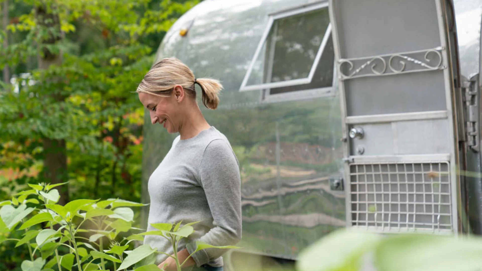 Woman smiling and looking at plants near a shiny trailer with an open door in a lush, green outdoor setting.