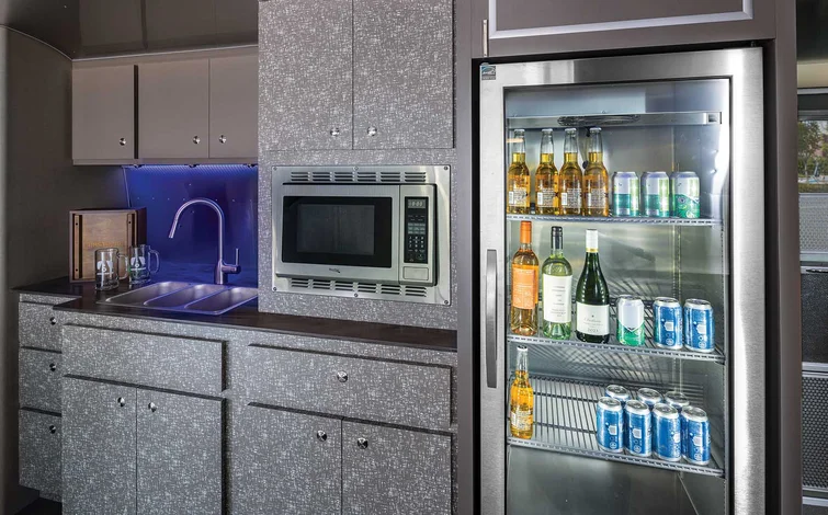 The interior of an Airstream Commercial custom bar and beverage trailer with sleek countertops, built-in microwave, stocked fridge, dual sinks, and overhead storage, featuring ambient ceiling lighting.