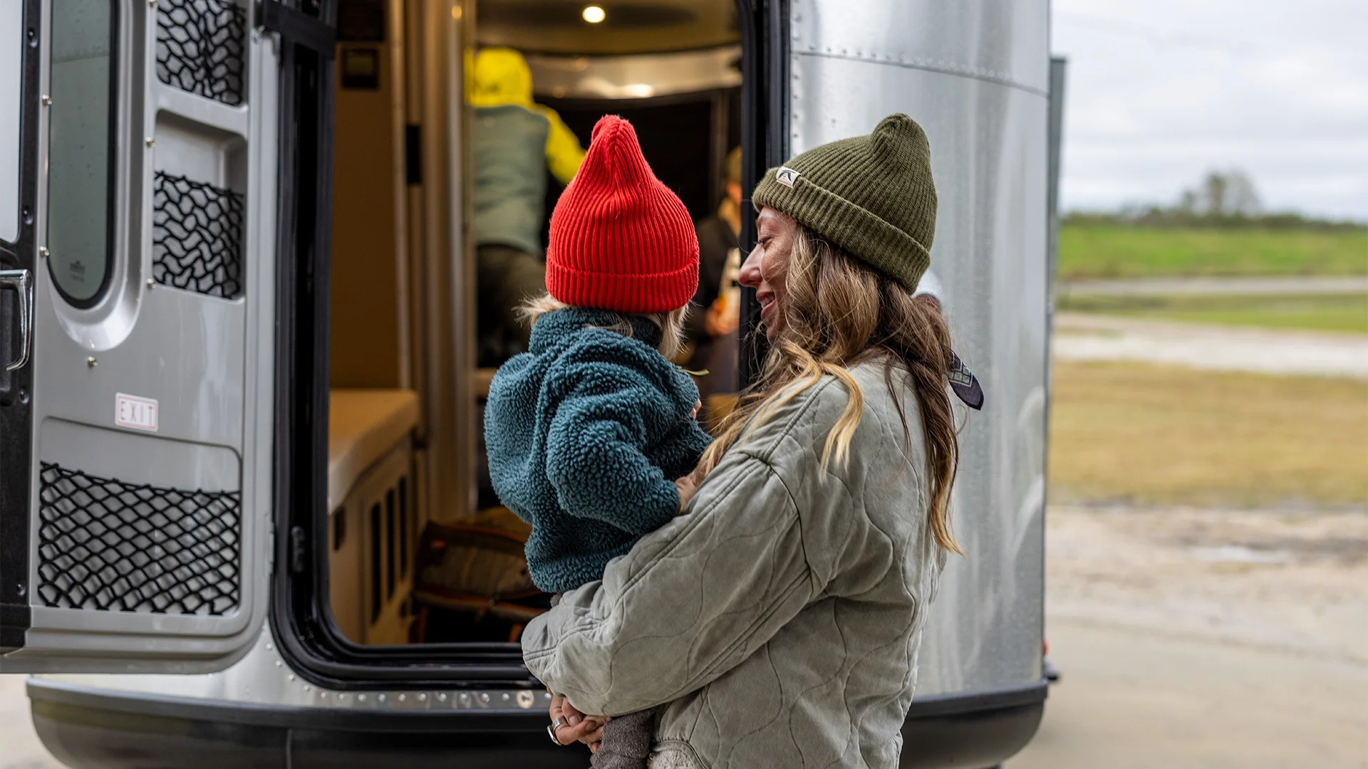 A woman in a green beanie holds a young child in a red hat near an open camper door, with a field in the background.