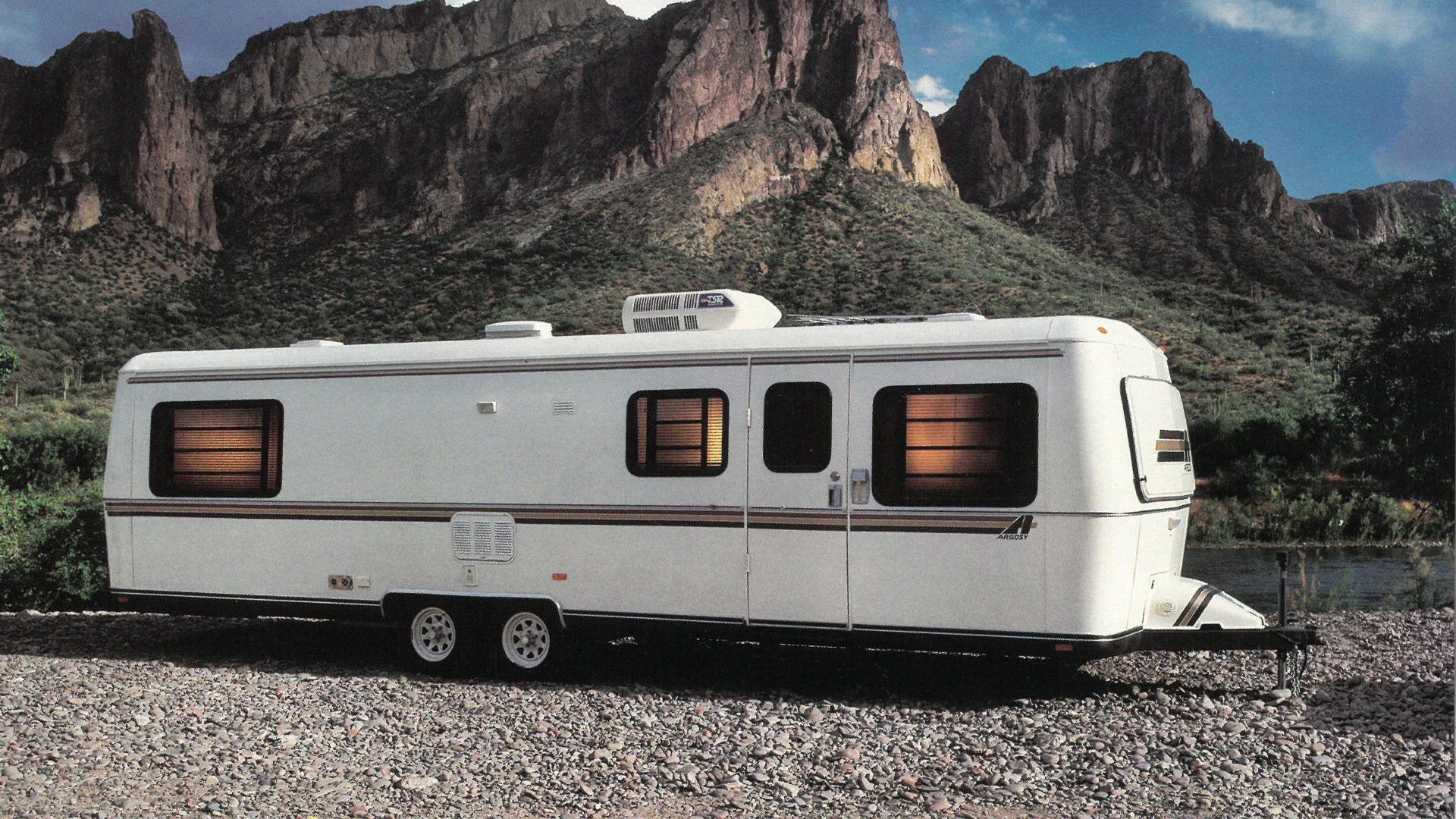 A white travel trailer with tinted windows is parked on gravel near a body of water, with tall, rugged mountains and a partly cloudy sky in the background.