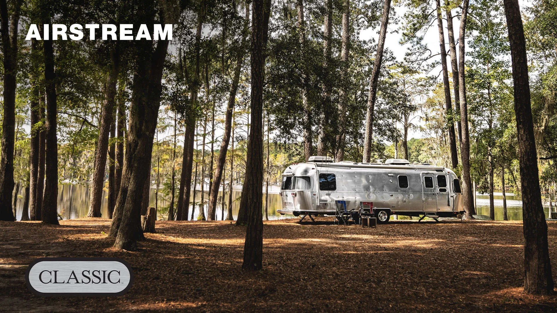 A silver Airstream trailer parked in a wooded area by a lake, surrounded by tall trees. Camping chairs and a table are set outside.