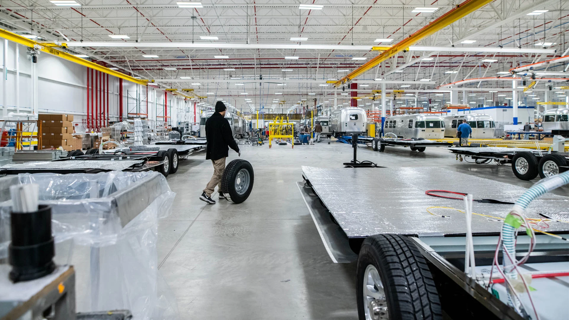 Person rolling a tire in a large, well-lit factory with trailers and equipment, featuring red and yellow overhead cranes.