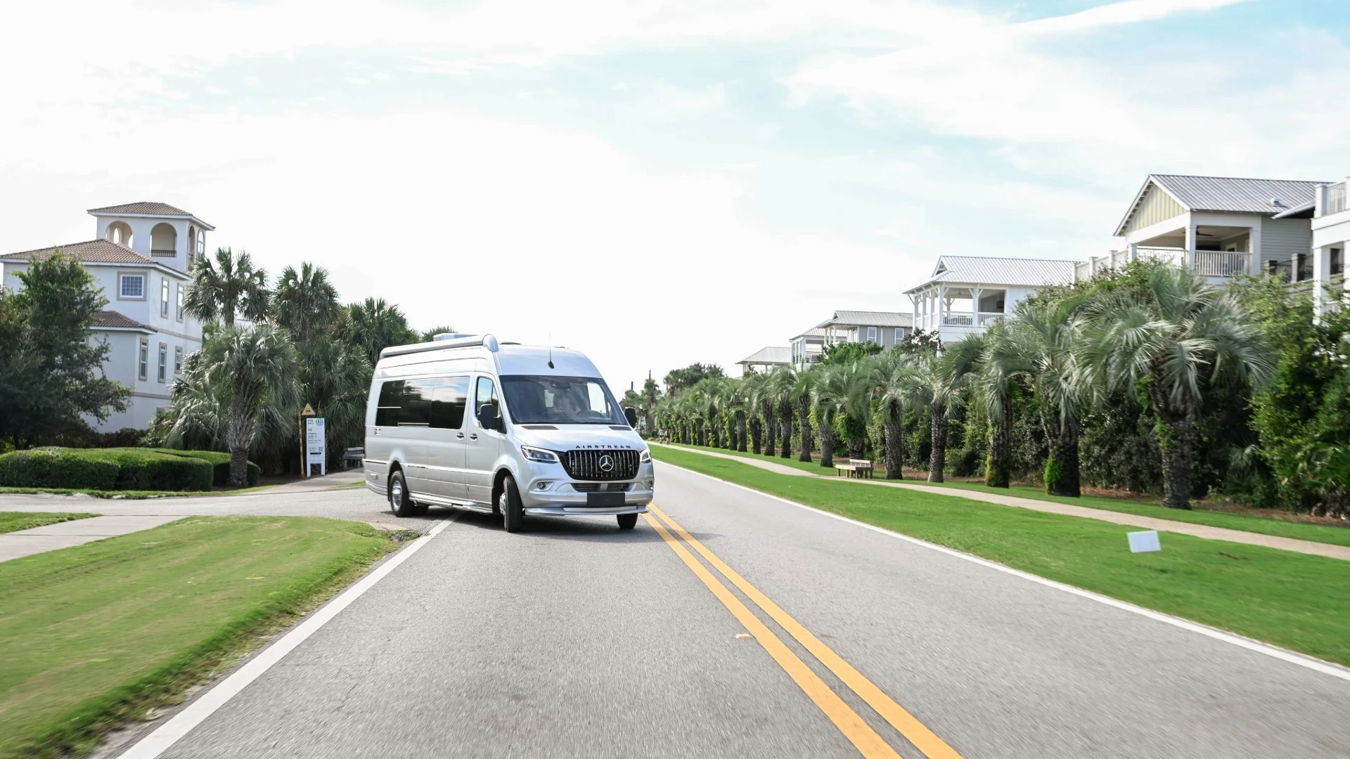 White van driving on a road lined with palm trees and modern houses under a clear sky.