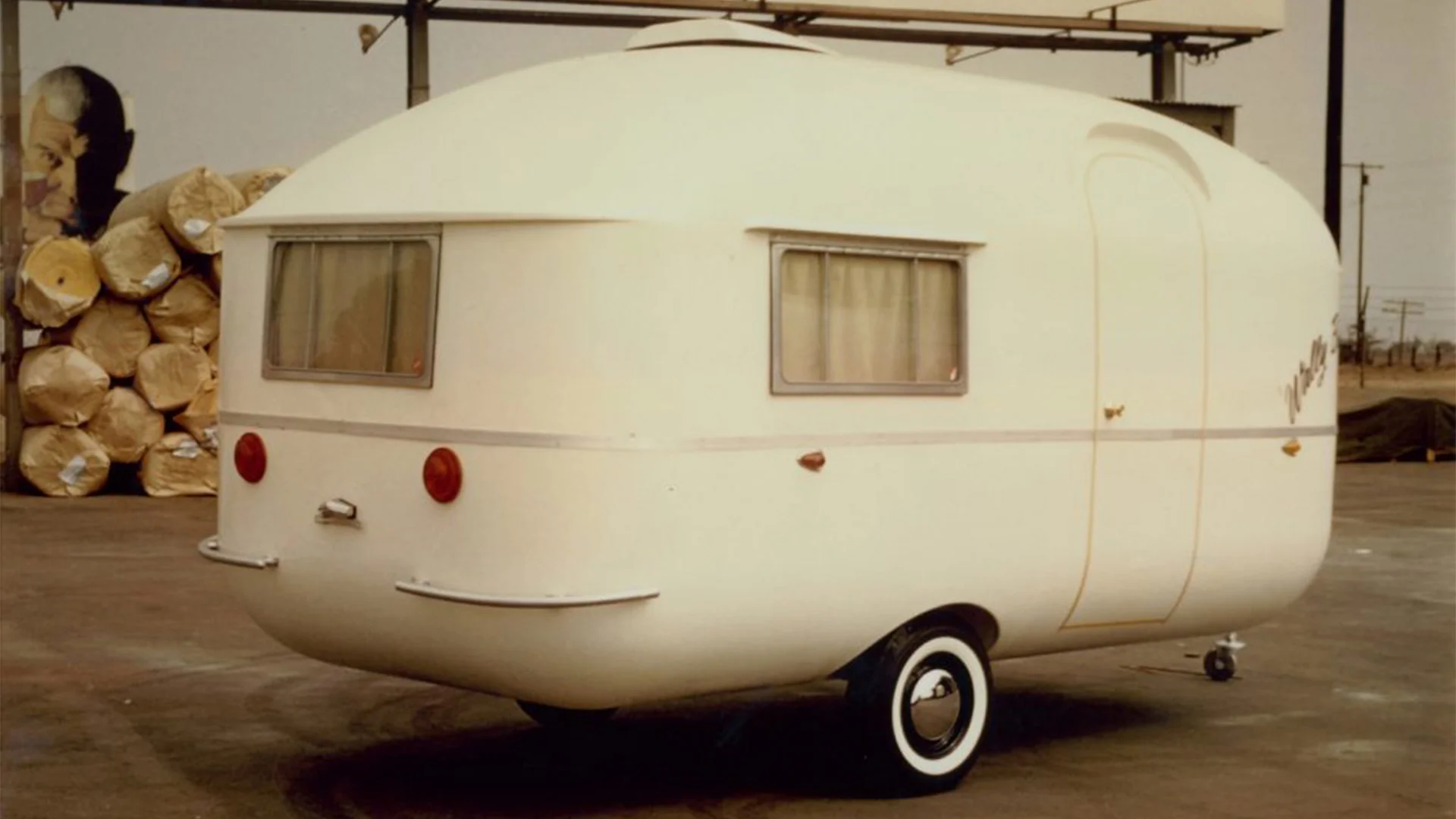 Vintage white teardrop-shaped trailer with small windows, parked indoors. The rear features round red taillights and a simple design.