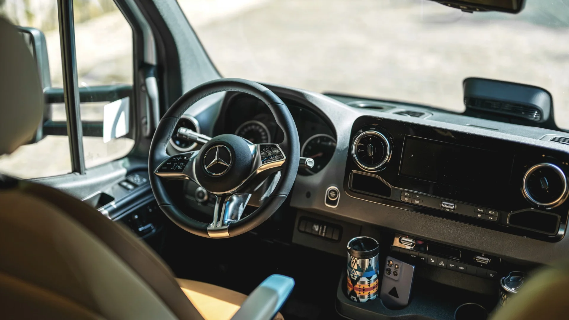 Interior of a Mercedes-Benz vehicle showing the steering wheel, dashboard controls, and cup holder with a patterned mug.
