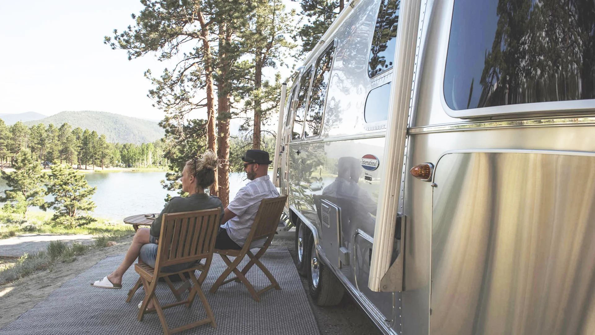 Two people sit on wooden chairs next to a shiny silver camper, enjoying a scenic view of a lake, pine trees, and distant mountains under a clear sky.
