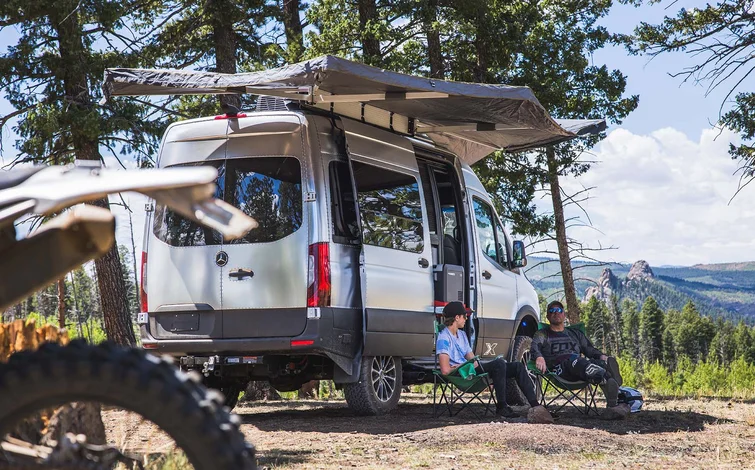 Two people relax in camping chairs under an awning attached to a silver van parked in a forested area, with mountains in the background and a motorcycle partially visible in the foreground.