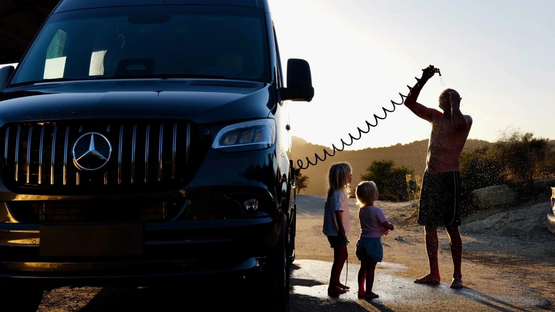 Man rinsing off with the exterior shower of an interstate camper van while two kids stand beside him