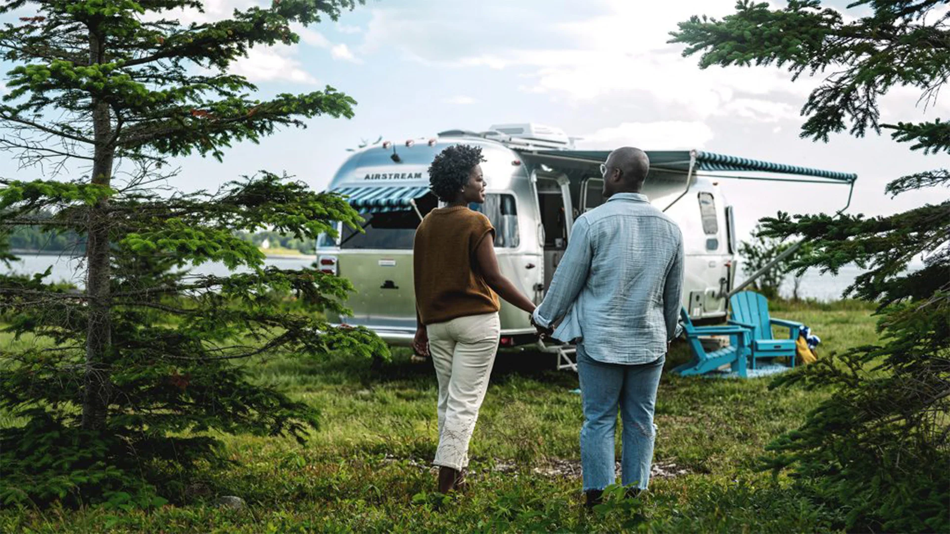 A couple holds hands in front of an Airstream trailer surrounded by trees, with Adirondack chairs nearby, under a partly cloudy sky.