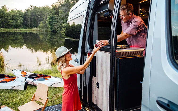 Woman in red dress receives drink from man in camper van near lake; kayaks and chair outside.