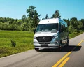 A silver camper van drives along a rural road with green fields and trees under a clear blue sky.