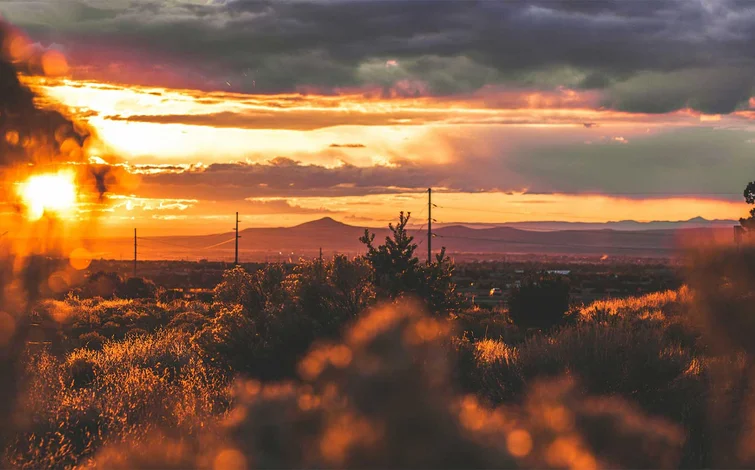 Sunset landscape with dramatic clouds, an orange sky, and distant mountains. Silhouetted trees and power lines in the foreground.