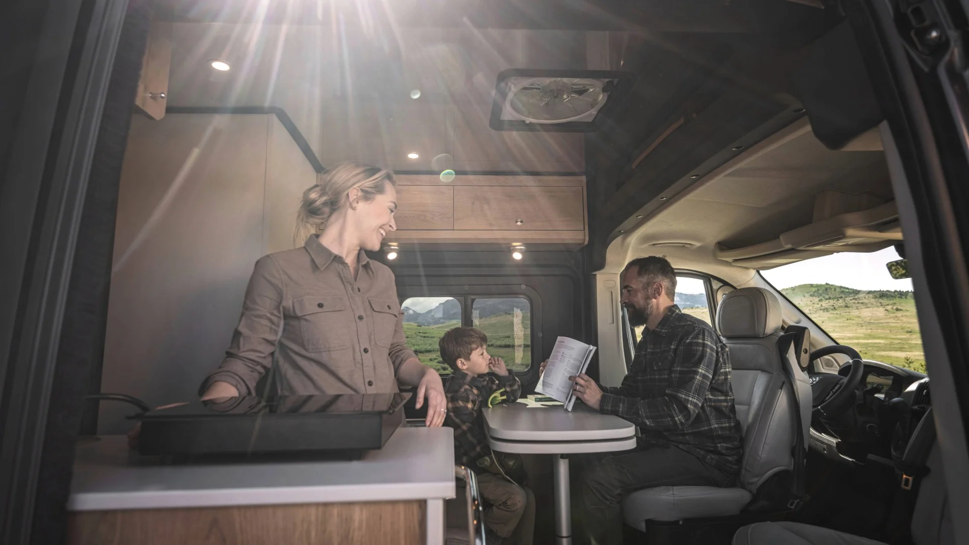 Family in a camper van, with a woman cooking and a man showing a book to a child, surrounded by nature under sunlight.