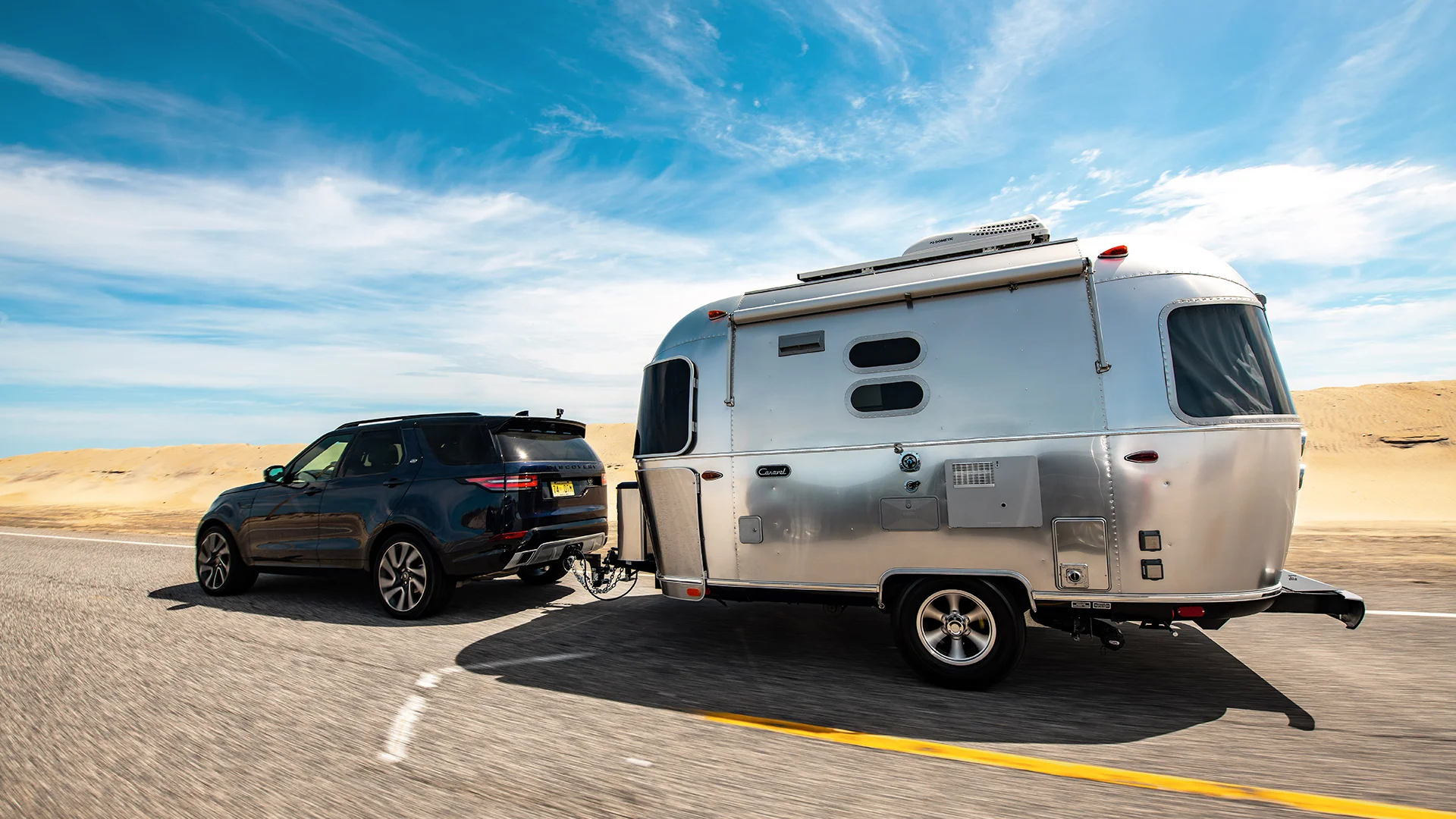 SUV towing a shiny silver caravan on a sunny desert highway, with clear blue skies overhead.