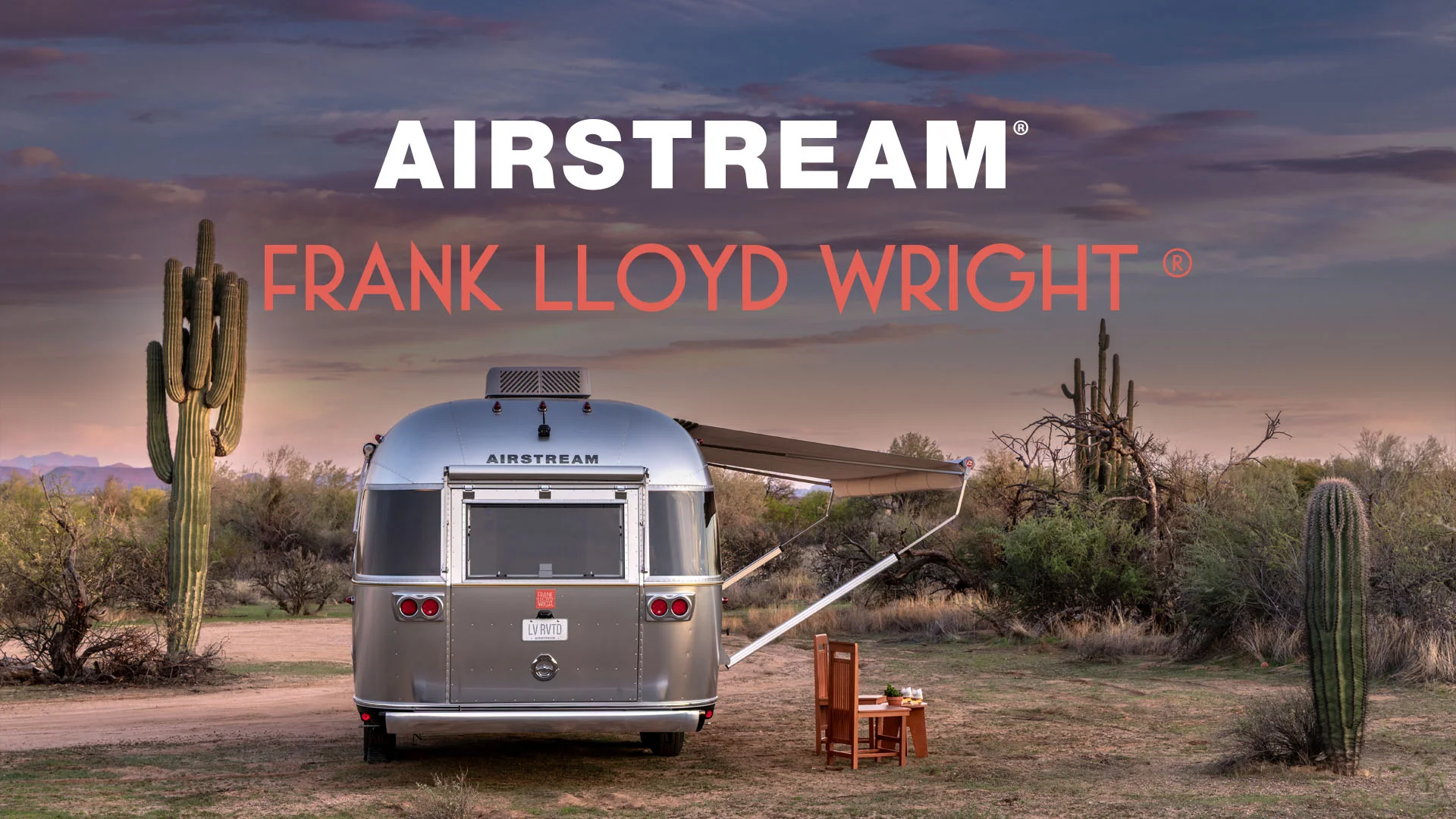 Airstream trailer with "Frank Lloyd Wright" text parked in a desert landscape, surrounded by cacti, under a colorful evening sky.