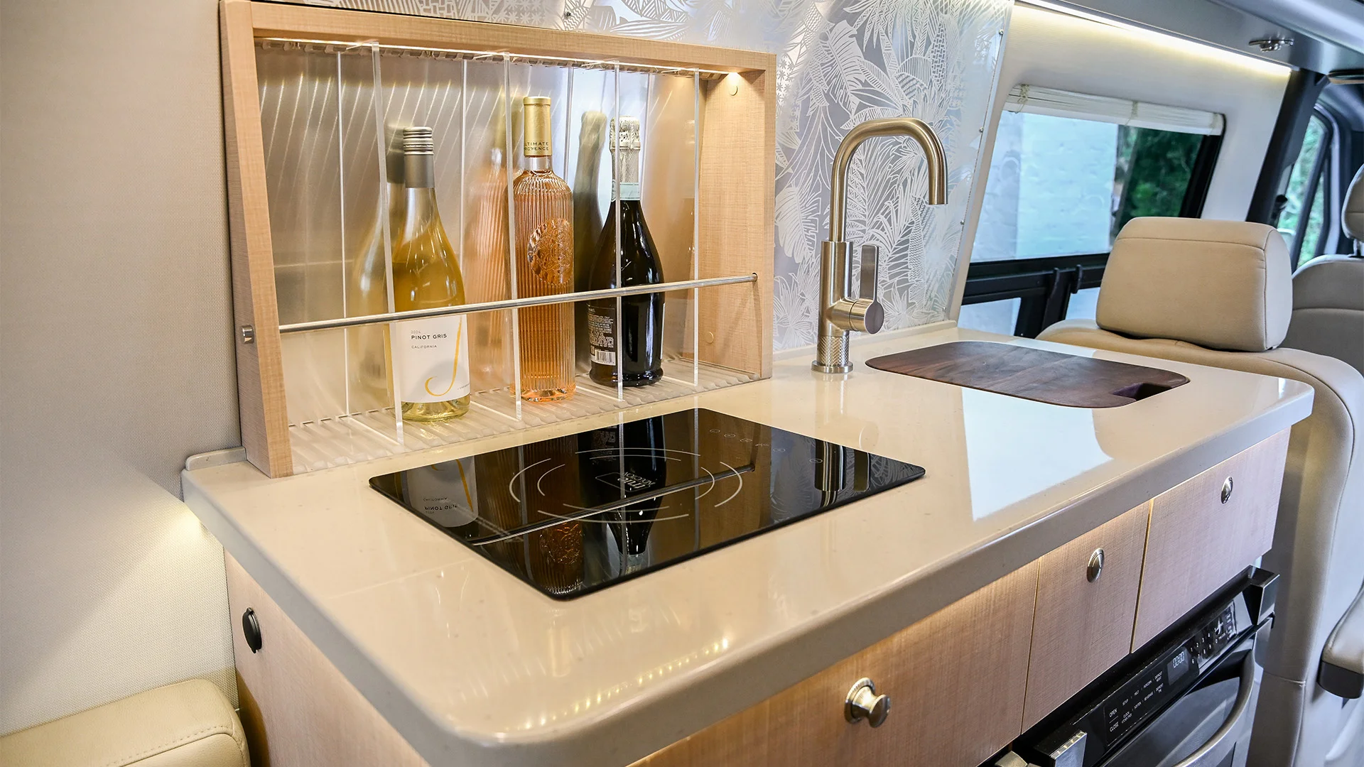 Modern RV kitchen with a glass-door cabinet holding wine bottles, an induction cooktop, stainless steel faucet, wooden countertop, and floral wallpaper.