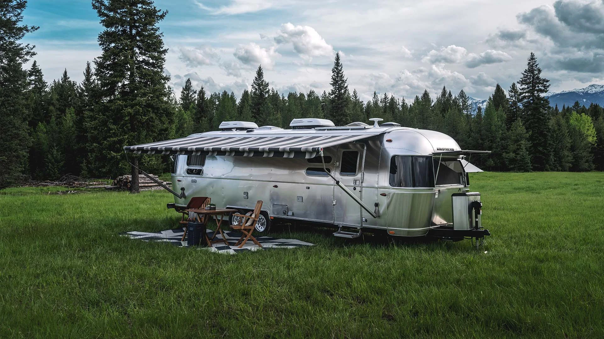 Airstream trailer in a grassy field with a striped awning extended, surrounded by trees, mountains, and cloudy sky. Folding chairs and table nearby.