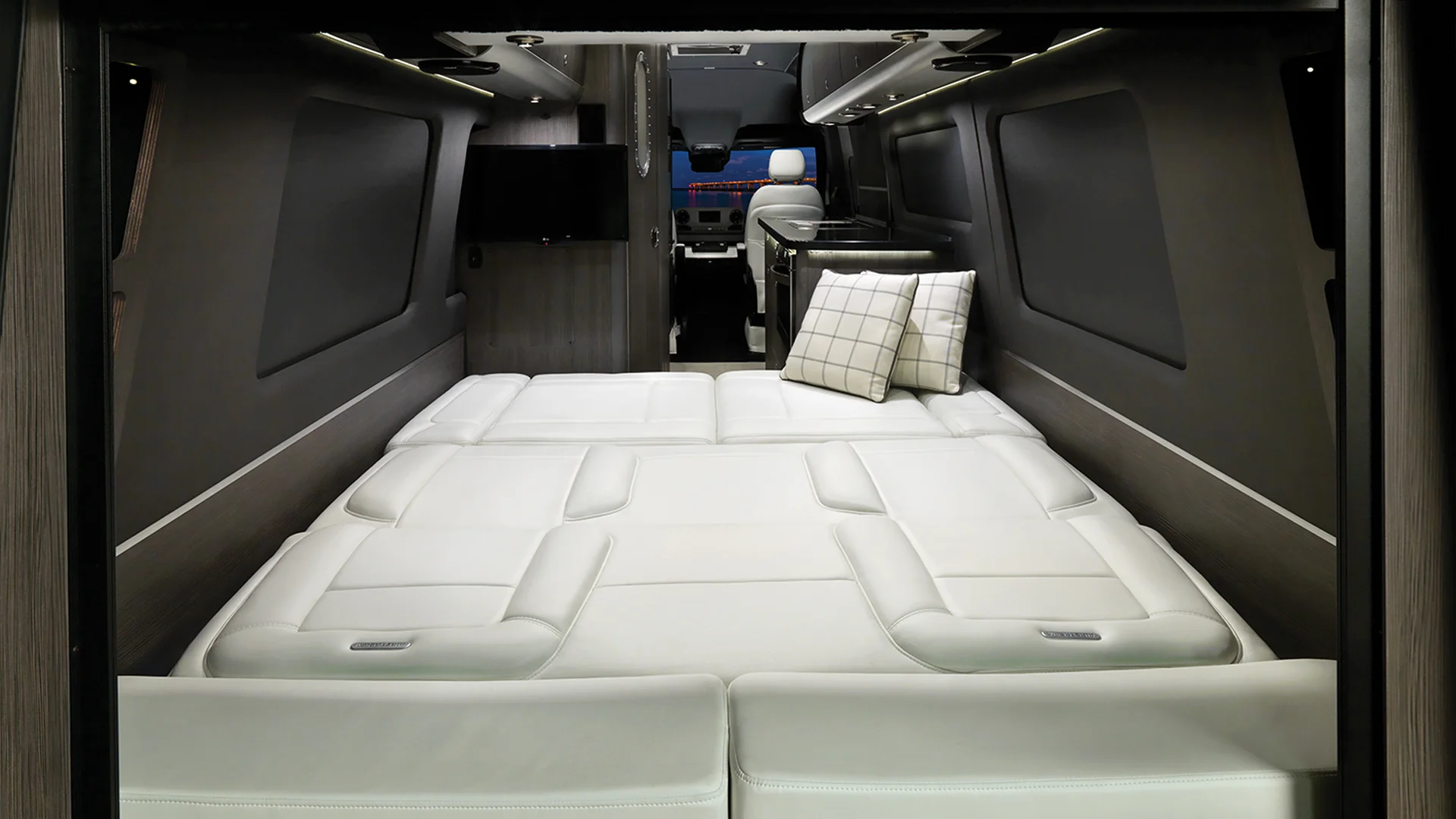 Luxury van interior featuring a spacious white bed with pillows, dark wood paneling, overhead storage, and a wall-mounted TV.