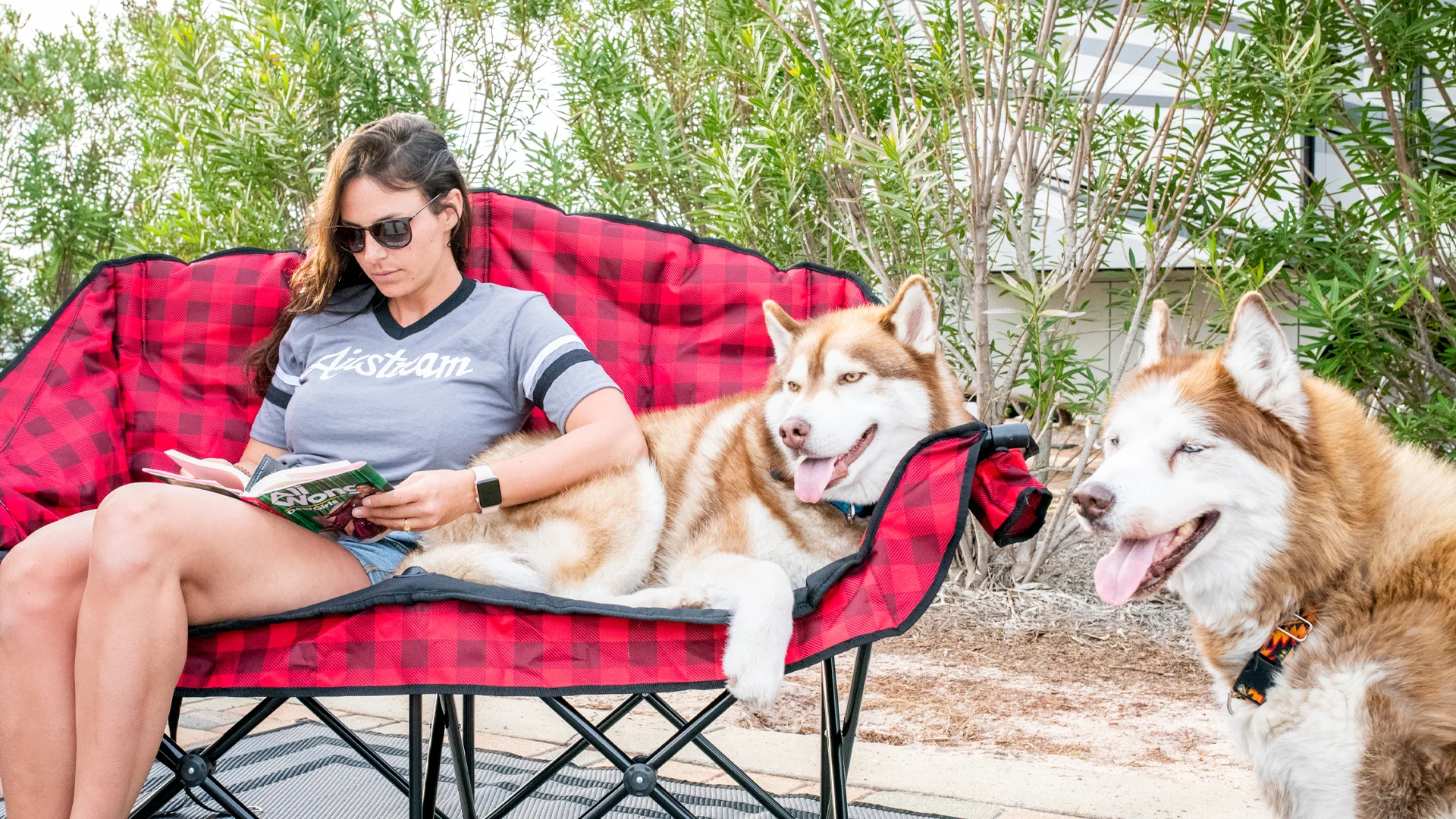Woman reading a book on a red plaid outdoor sofa with two Siberian Huskies, surrounded by greenery.