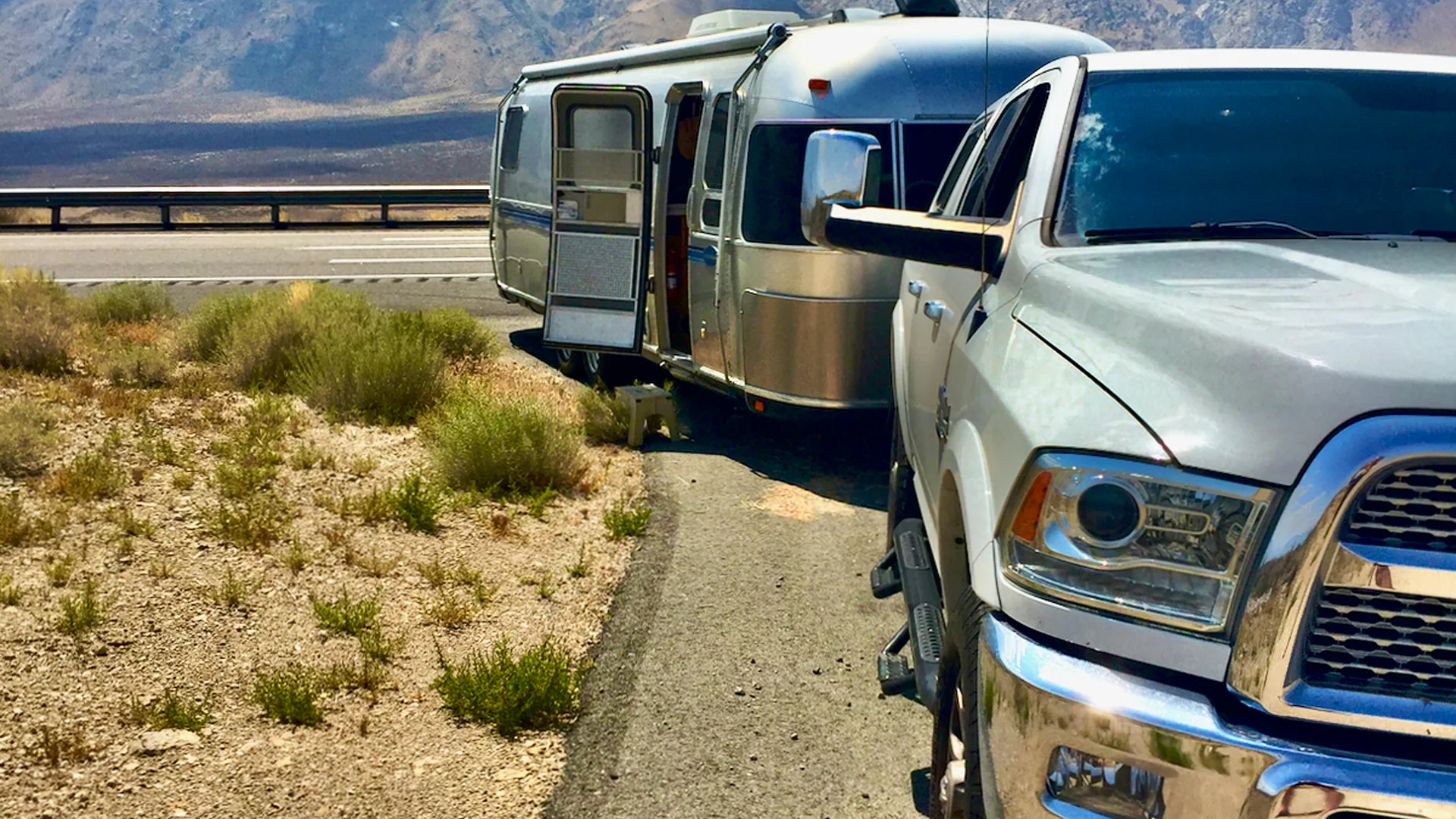 A silver pickup truck towing an Airstream trailer is parked on the side of a road near dry grassland and mountains, with the trailer door open.