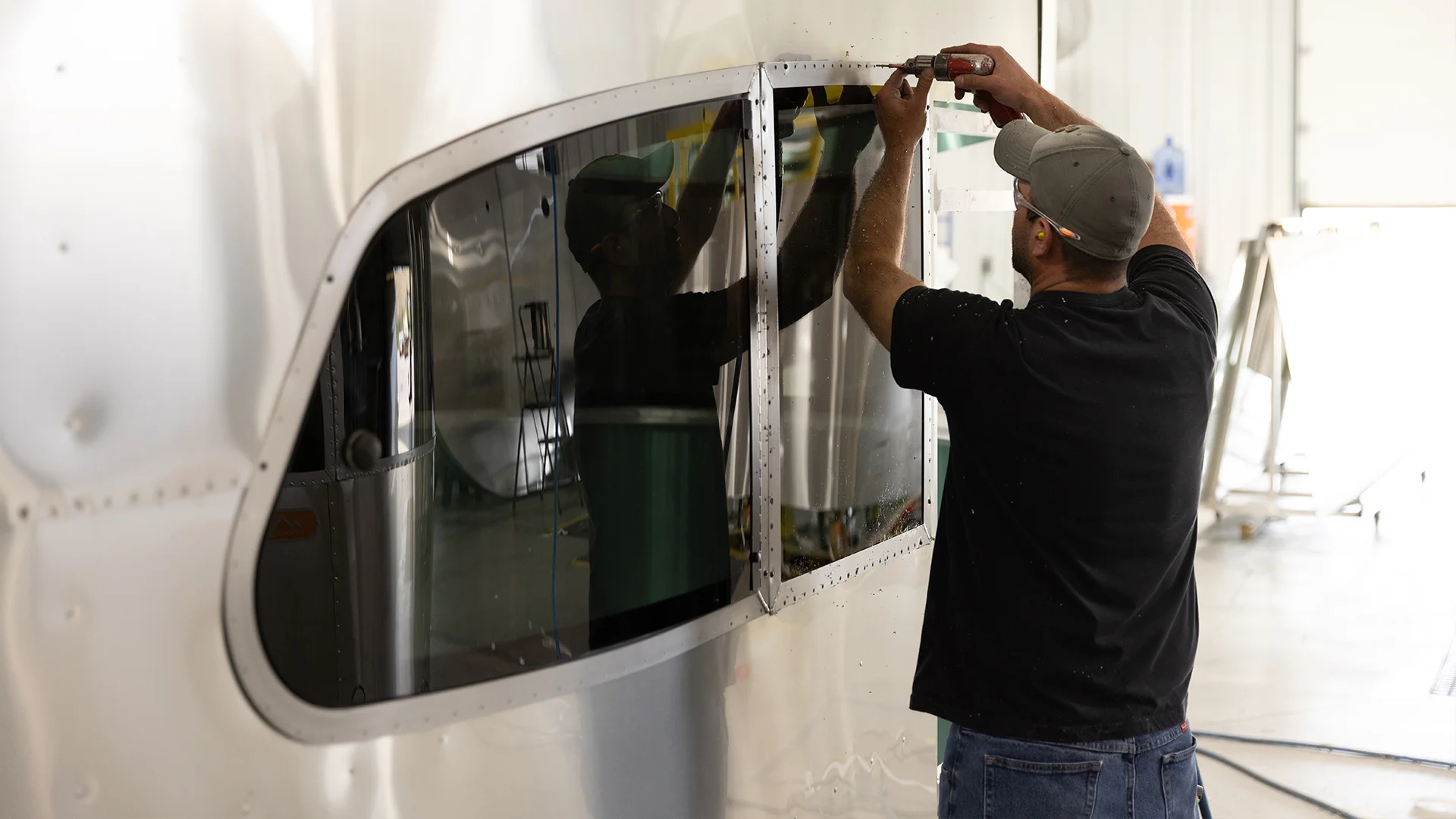Man in a black shirt working on a window frame of a shiny silver trailer, reflected in the surface, inside a well-lit workshop.