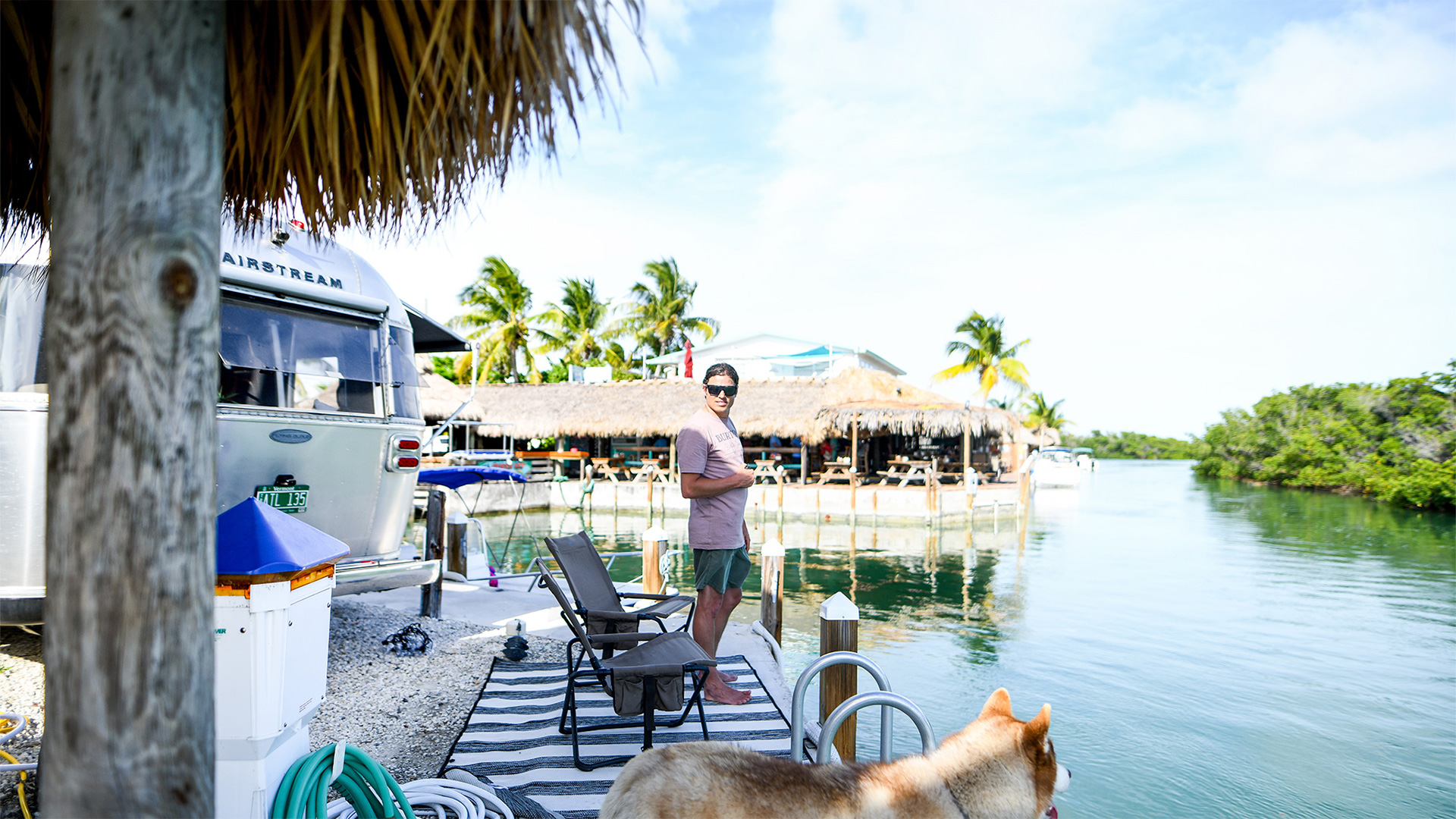 Man standing near an Airstream trailer by the water, with palm trees, a dog, chairs, and a thatched hut in the background.