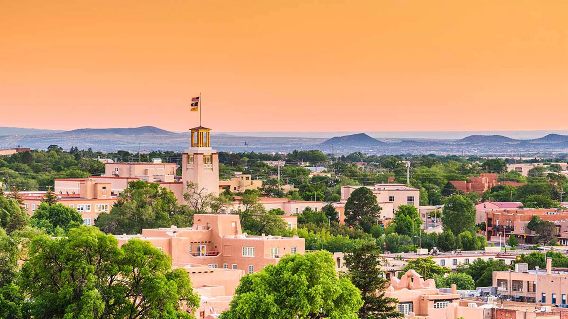 Cityscape with adobe-style buildings, lush trees, and a tower flying a flag against a vibrant orange sky, set against distant hills.