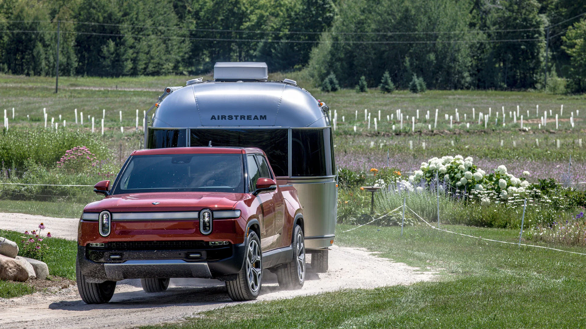 A red electric pickup truck towing a silver Airstream trailer on a dirt road through a grassy area with flowers and trees in the background.