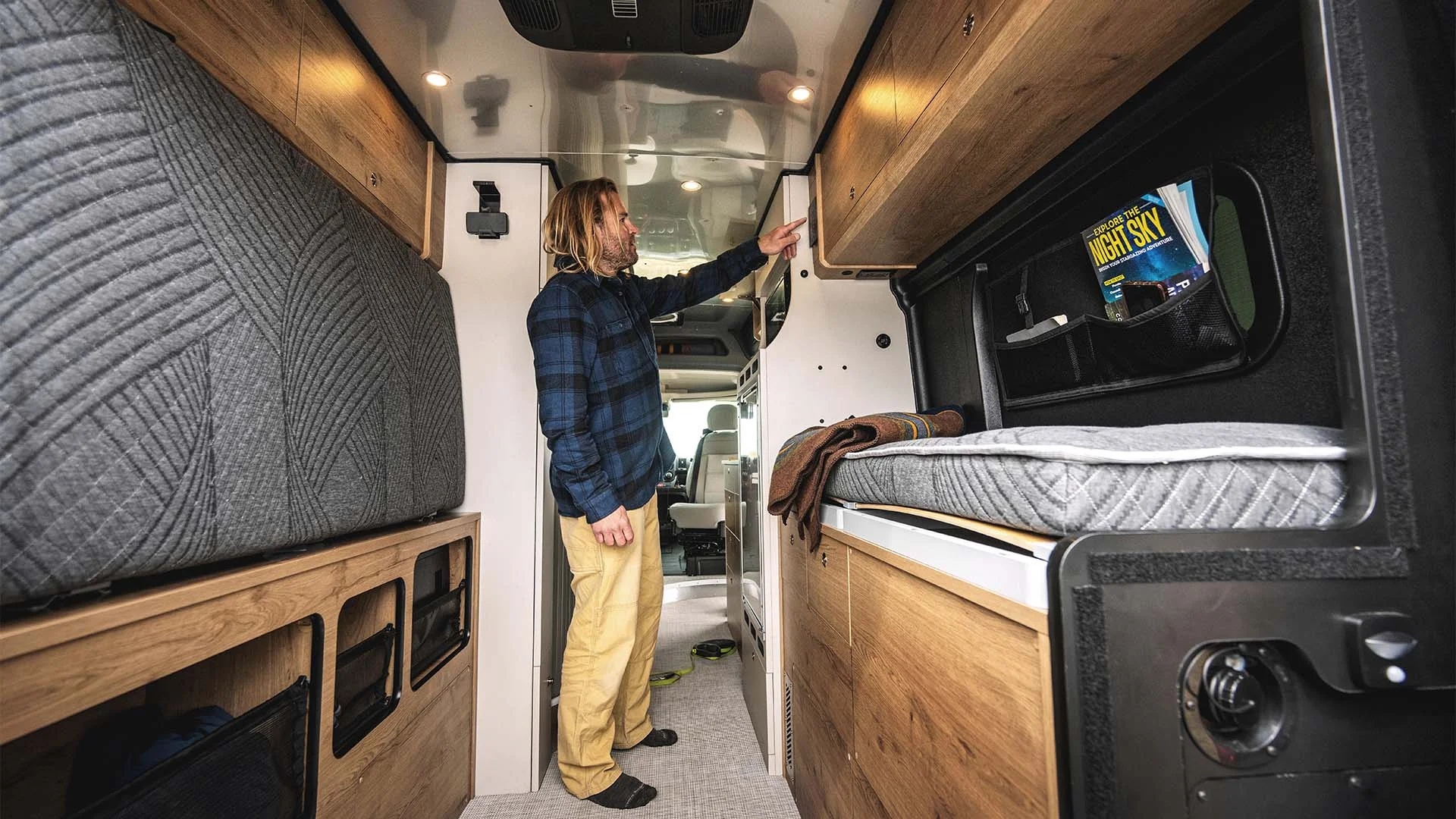 Person in a flannel shirt checking overhead storage in a cozy, wood-accented camper van interior.