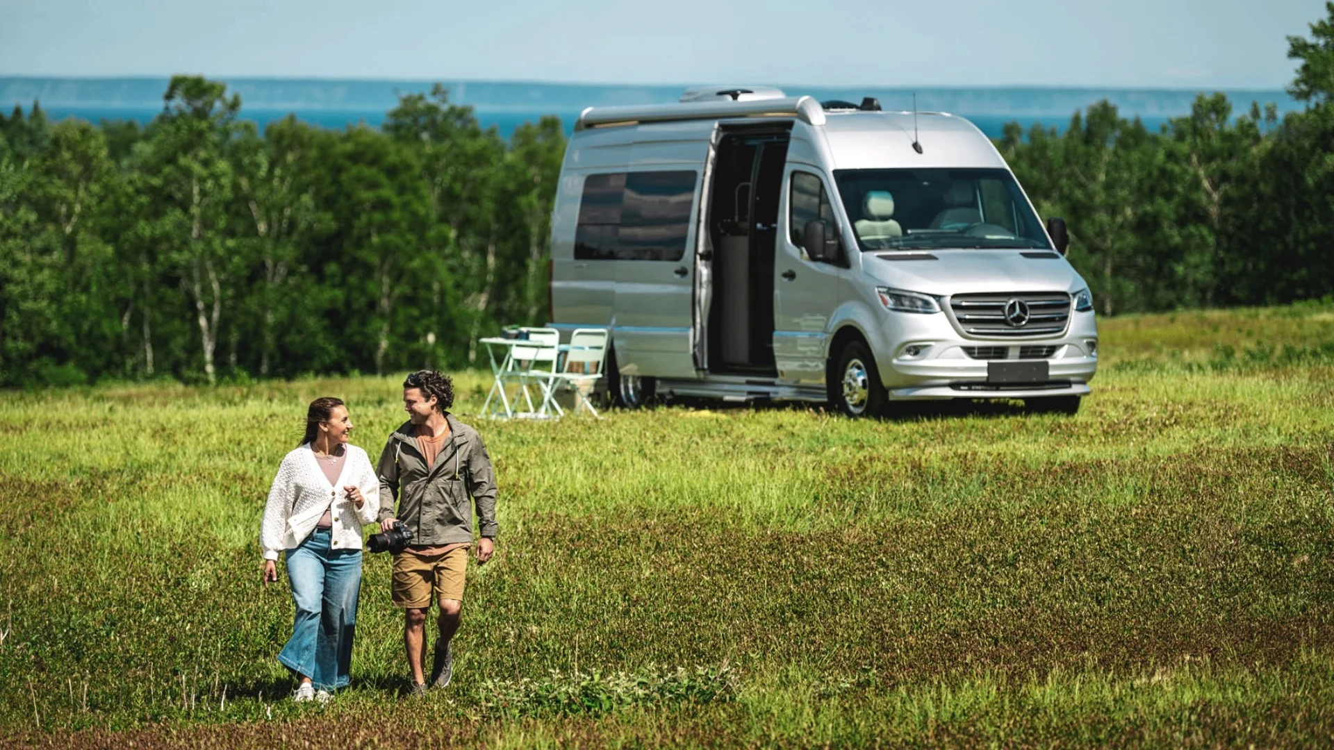 A couple walks in a grassy field near a parked silver camper van with a folded table and chairs, surrounded by trees and a distant view of water.