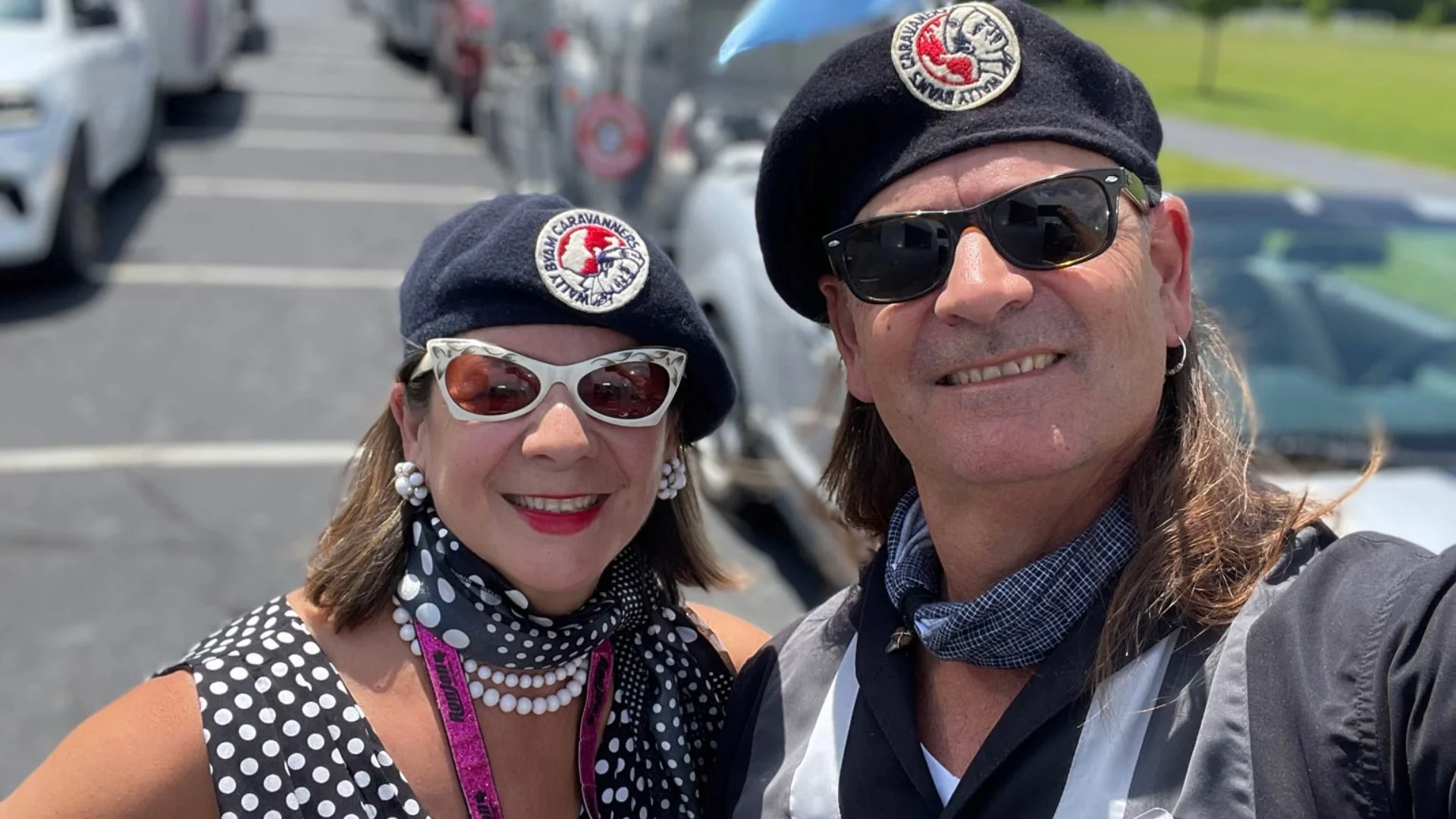 Two people wearing black berets and sunglasses, standing outdoors. The woman wears polka dots and pearls, the man in a black scarf and jacket.