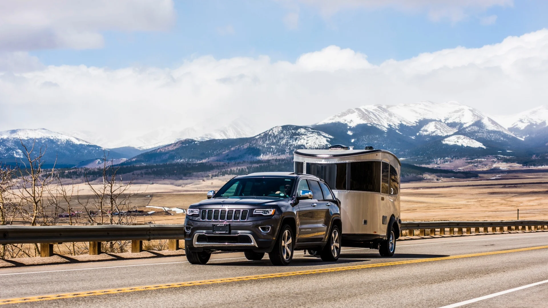 SUV towing a camper on a highway with snow-capped mountains and a vast plain in the background.