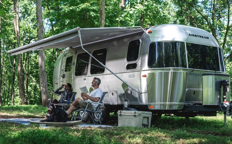 A couple relaxes under the awning of a shiny Airstream trailer in a lush forest; one reads a book, the other uses binoculars.