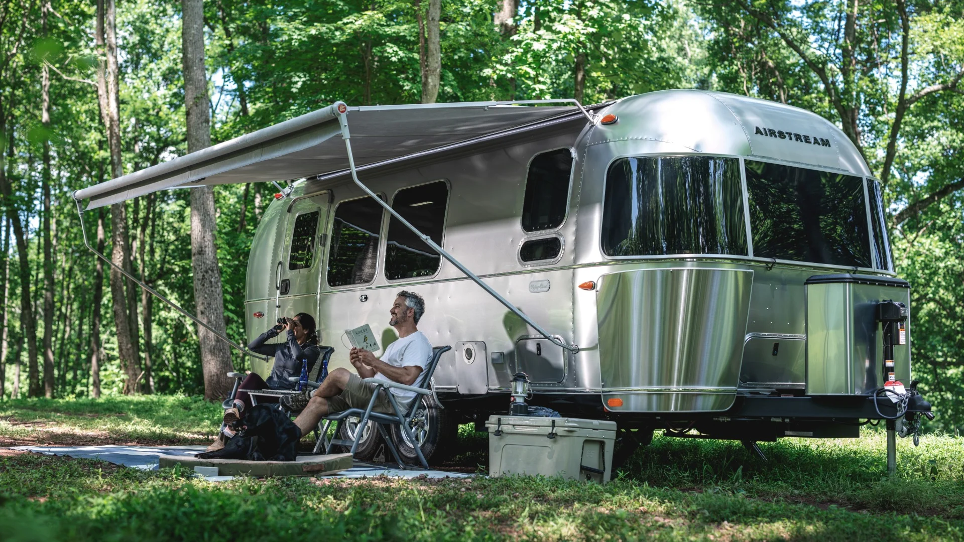 A couple relaxes under the awning of a shiny Airstream trailer in a lush forest; one reads a book, the other uses binoculars.