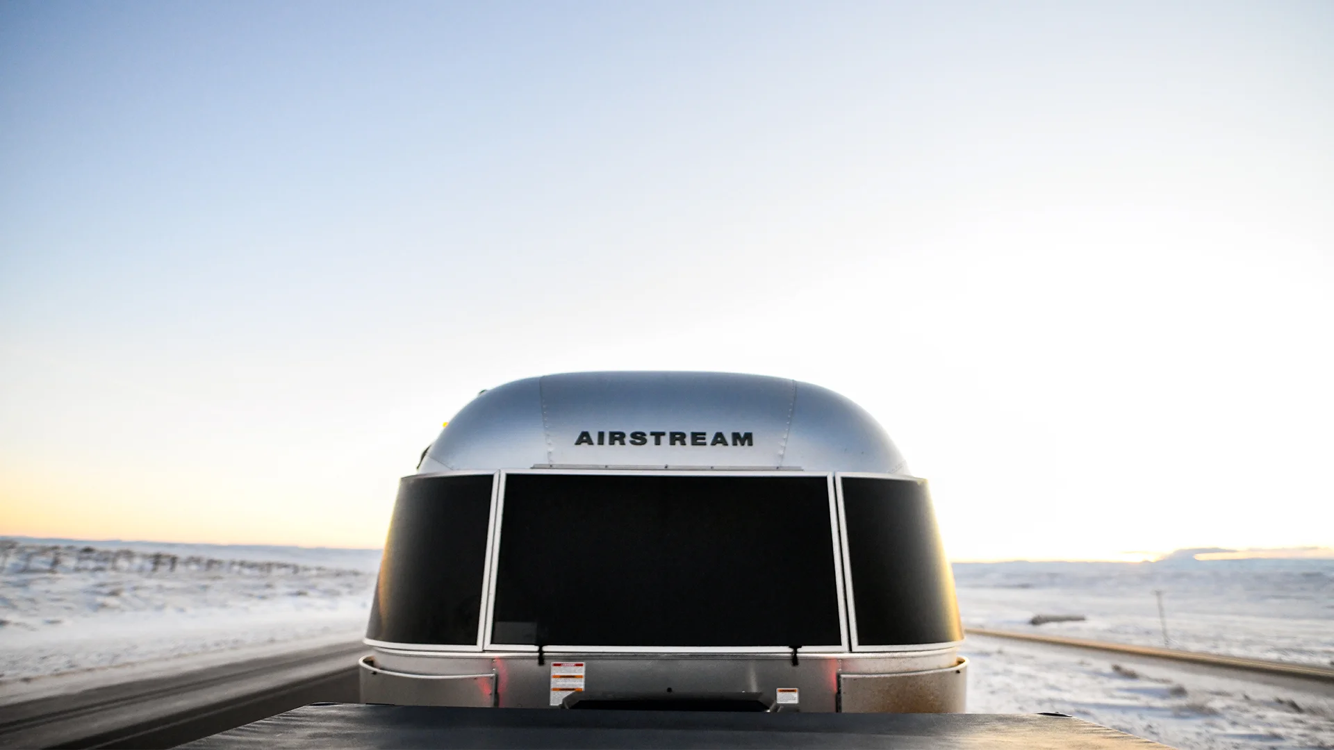 A silver Airstream trailer on a snowy road under a clear sky, viewed from the back with the horizon in the distance.
